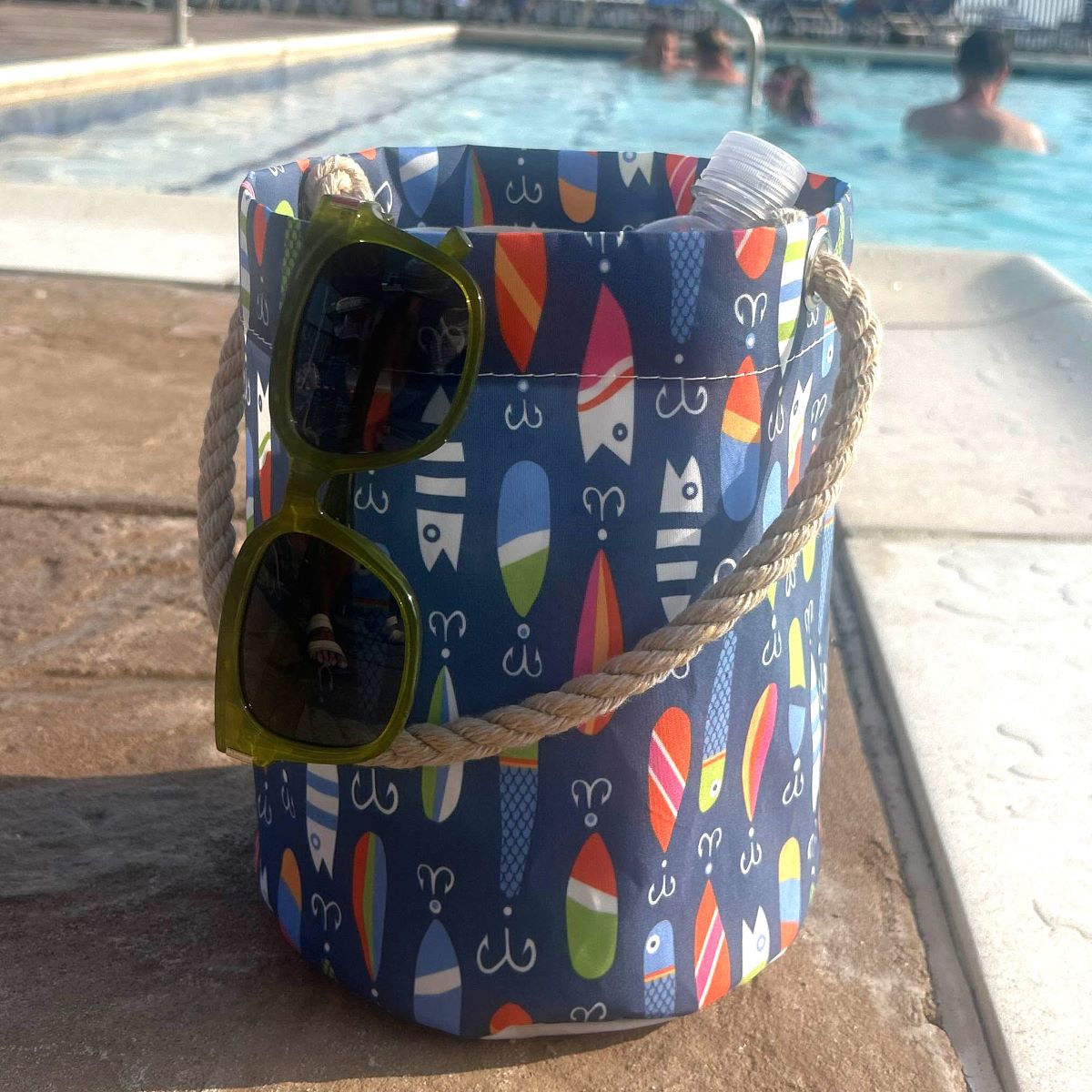 A Kate Nelligan Lures Bucket Bag by Sea Bags, featuring a blue design with surfboard motifs and a sturdy rope handle, holds a water bottle and sunglasses as it rests poolside—an essential nautical accessory adorned with coastal-inspired prints. by Sea Bags