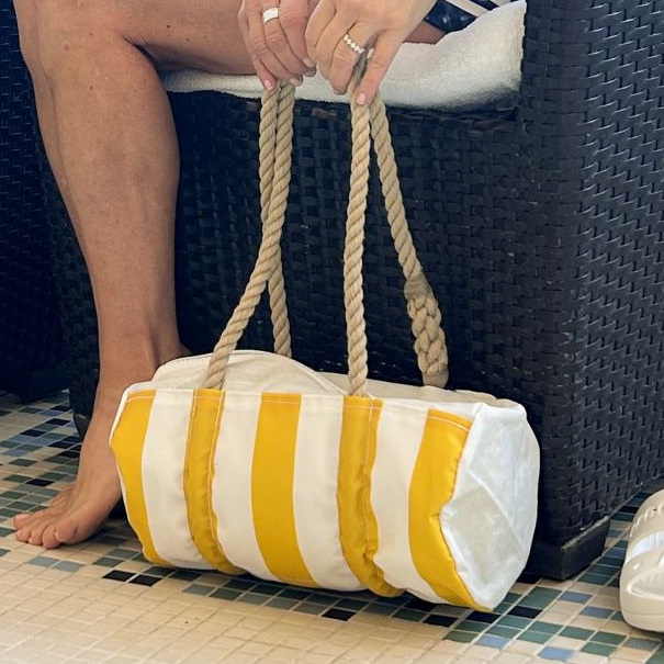 A person sits on a wicker chair, holding the Sea Bags Sun  Sea Stripe Baguette Bag with rope handles. Their feet are bare, and the floor features a mosaic tile pattern. by Sea Bags
