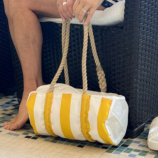 A person sits on a wicker chair, holding the Sea Bags Sun  Sea Stripe Baguette Bag with rope handles. Their feet are bare, and the floor features a mosaic tile pattern. by Sea Bags