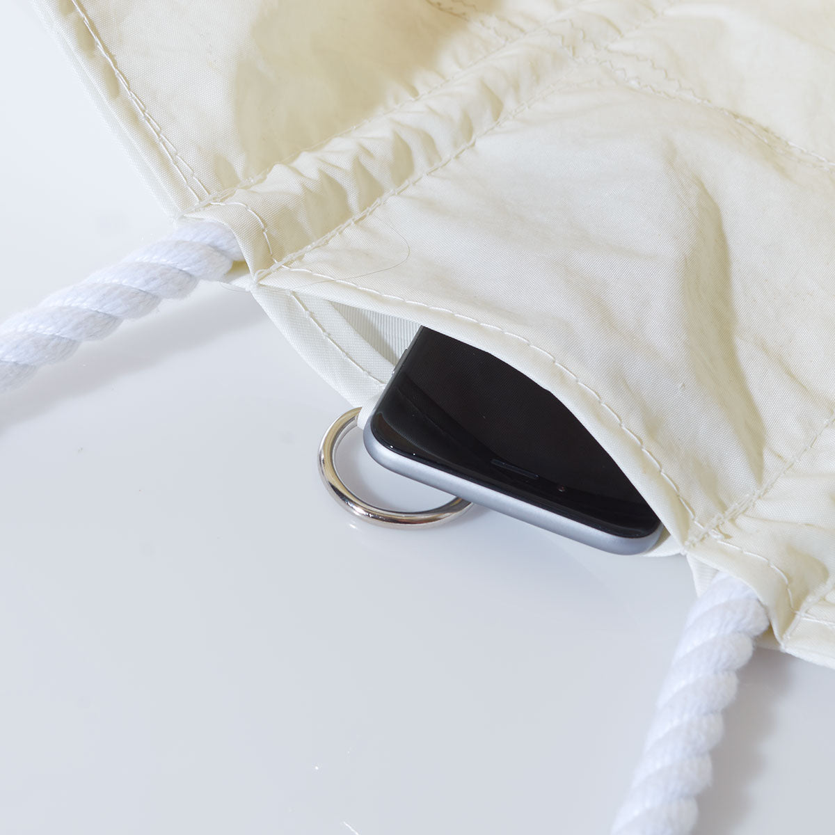 A smartphone and a keyring are partially visible inside the Sea Bags Custom Design Medium Tote, a beige reclaimed sail bag with white rope handles, placed on a white surface. by Sea Bags