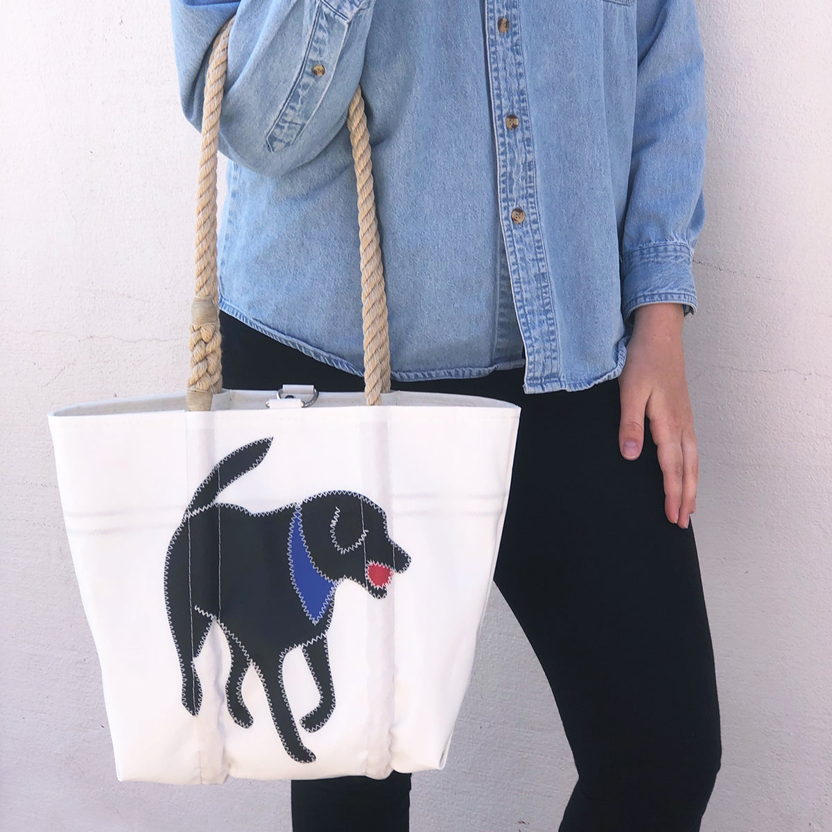 A person dressed in a denim shirt and black pants holds the Sea Bags Custom Design Handbag—a white tote featuring a black dog motif, crafted from reclaimed sail bag material and finished with sturdy rope handles. by Sea Bags
