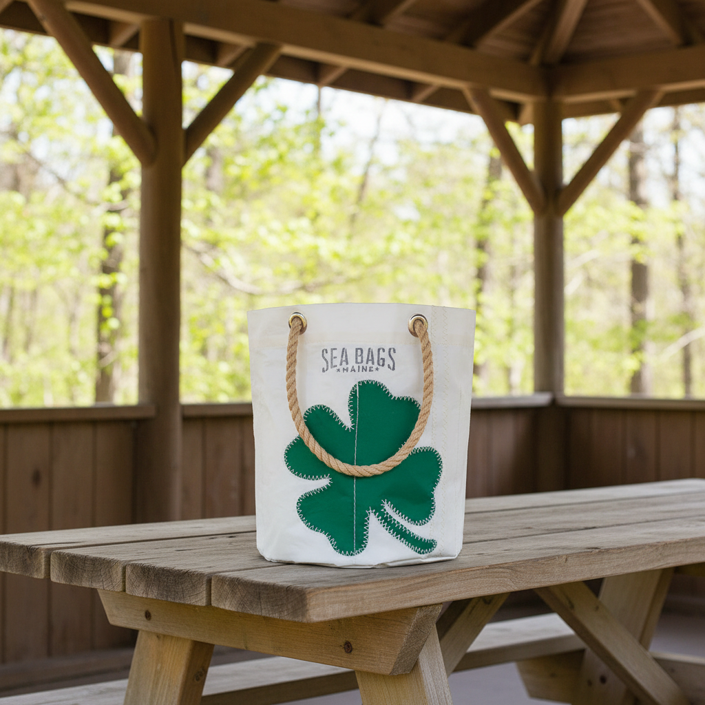 The Four Leaf Clover Beverage Bucket Bag by Sea Bags, crafted from recycled sail cloth with rope handles and adorned with a green shamrock design, rests on a wooden picnic table under a gazebo in a wooded area. by Sea Bags