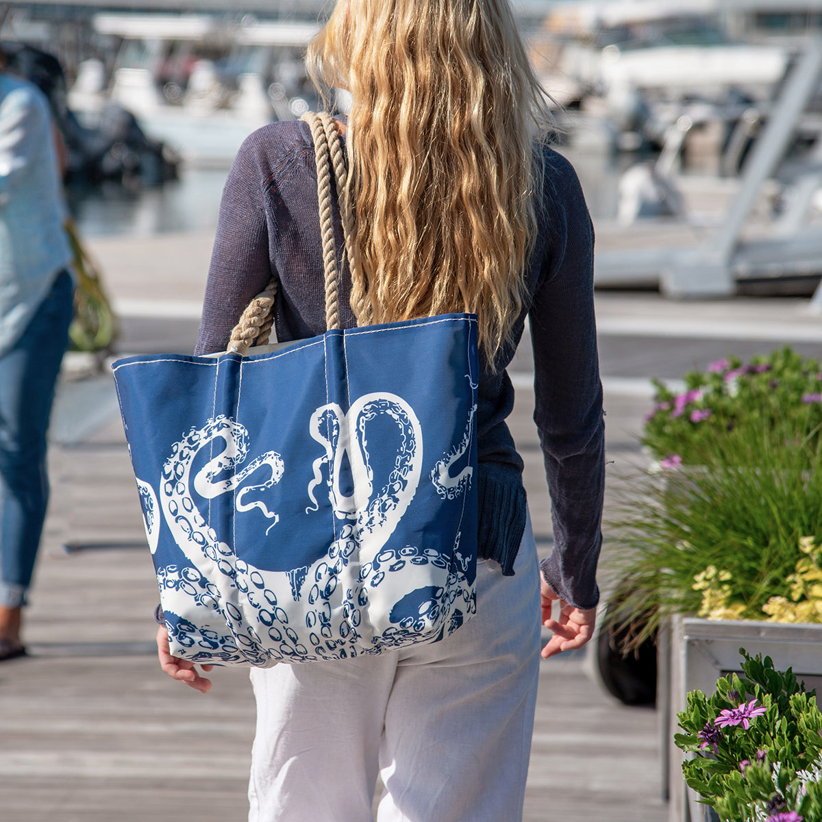 A person walks on a dock carrying the Sea Bags Zip Top White on Navy Octopus Large Tote, featuring hand-spliced rope handles and crafted from recycled sail cloth; plants and boats can be seen in the background. by Sea Bags