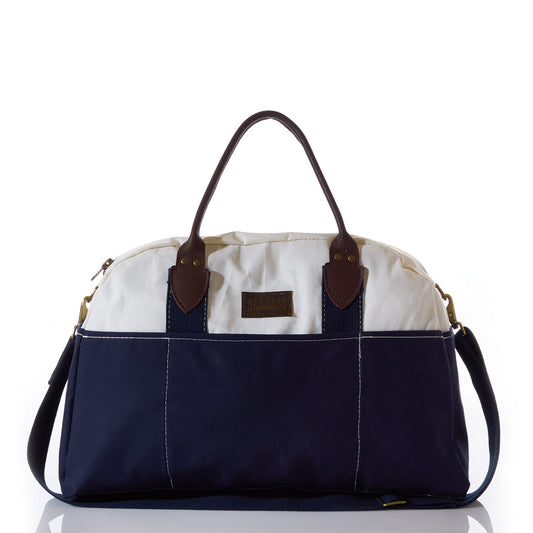 The Chebeague Weekender by Sea Bags is a two-tone weekender bag made from recycled sail cloth, featuring a white top, dark blue bottom, brown leather handles, a shoulder strap, and a small front label—all displayed against a white background. by Sea Bags