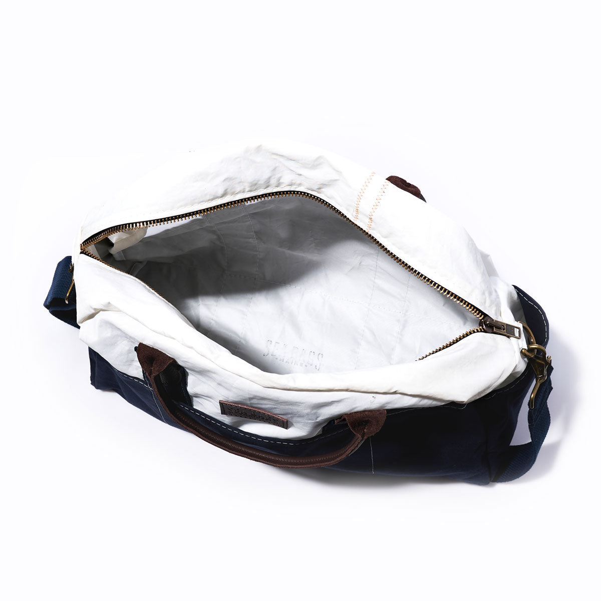 Top view of the Chebeague Weekender by Sea Bags, featuring white lining, brown leather handles, a blue body made from recycled sail cloth, and a brass zipper. The empty bag rests on a white background. by Sea Bags