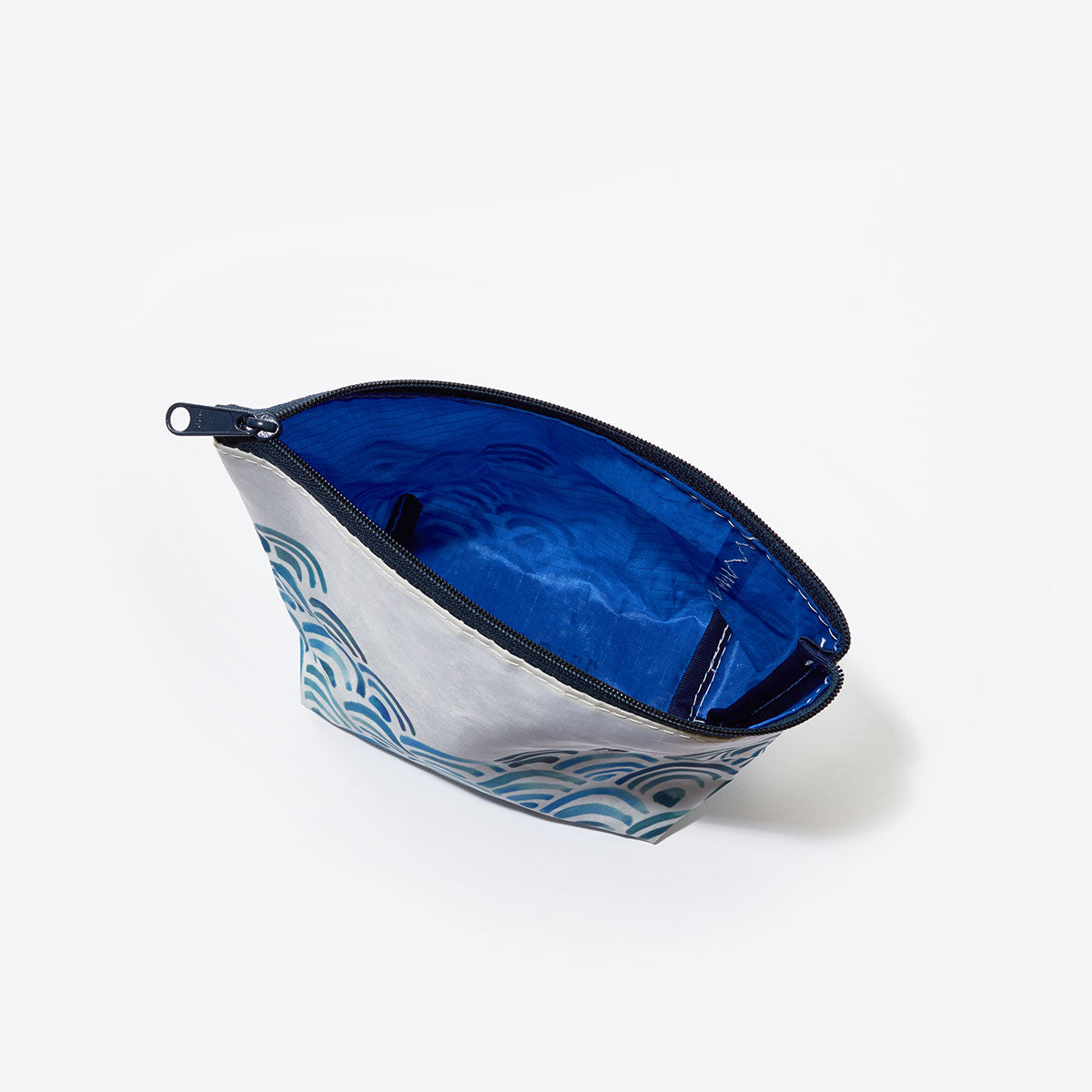 The Sea Bags Watercolor Waves Cosmetic Bag features a blue wave pattern on the exterior and a blue interior. Shown open from a top angle against a white background, this small zippered pouch is crafted from recycled sailcloth, making it a stylish and eco-friendly travel accessory. by Sea Bags