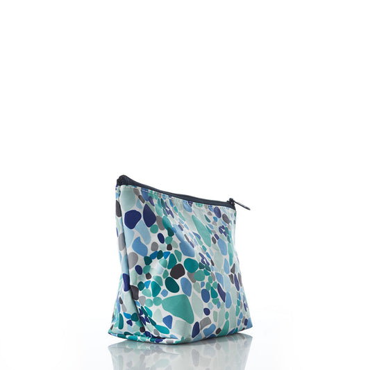 The Sea Bags Sea Glass Print Cosmetic Bag sits upright on a white reflective surface, showcasing a fabric zipper pouch adorned with an abstract pebble pattern in shades of blue, teal, and rare blue reminiscent of sea glass. by Sea Bags