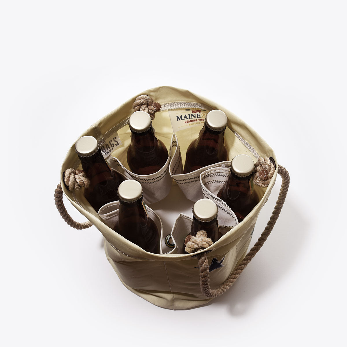 Top view of the Maine Bicentennial Beverage Bucket by Sea Bags, a tan tote crafted from recycled sail cloth with rope handles. Featuring individual compartments that securely hold six brown glass bottles and space for a book or pamphlet, this versatile bag captures classic maritime style and draws inspiration from the spirit of Maine’s 1901 state flag. by Sea Bags
