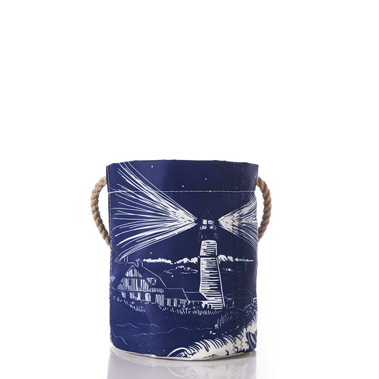 Introducing the Lighthouse Beacon Bucket Bag by Sea Bags—a navy blue recycled sail cloth bucket tote with rope handles, adorned with a white illustrated Maine lighthouse and coastal scene reminiscent of Portland Head Light. This sustainable accessory seamlessly blends coastal charm with eco-conscious craftsmanship. by Sea Bags