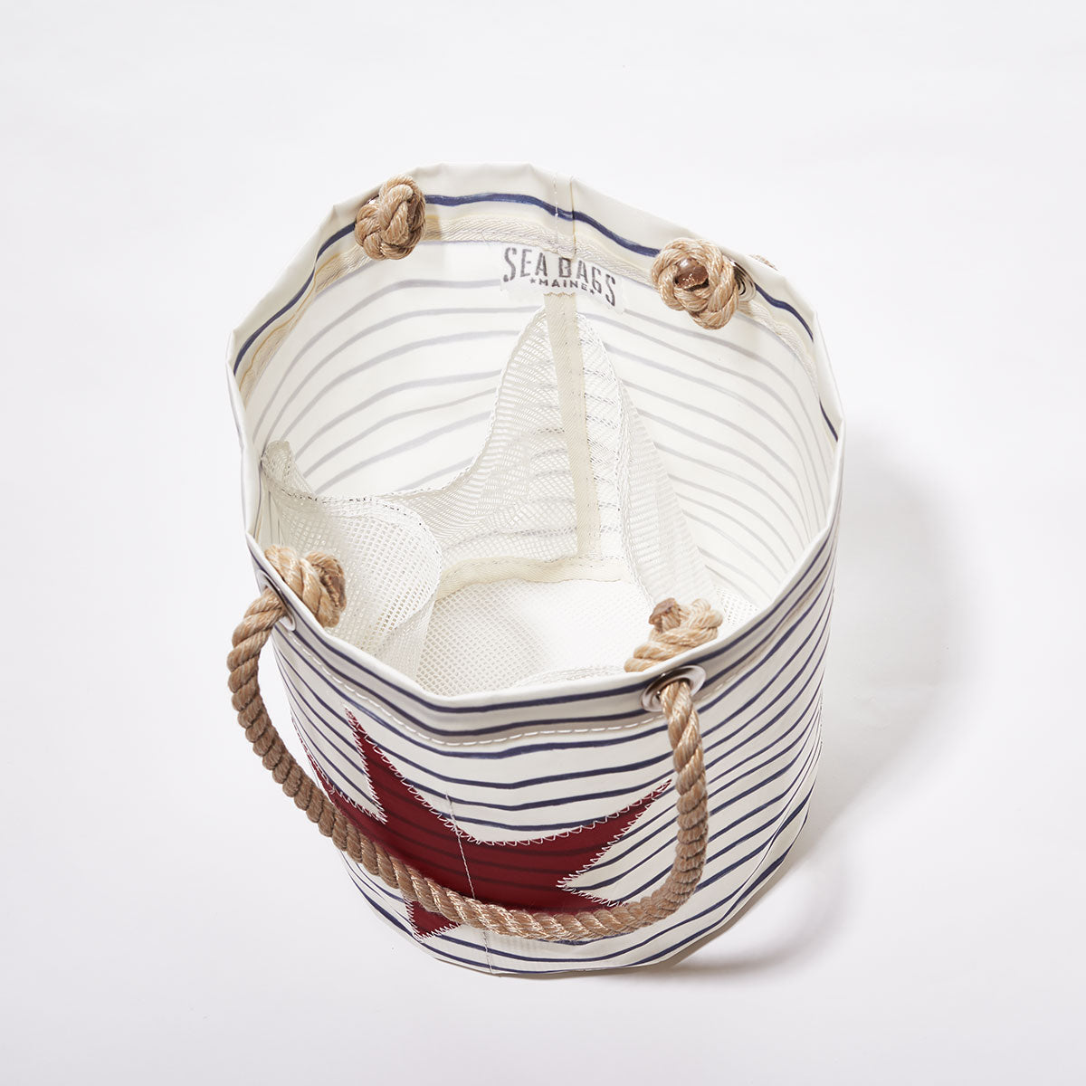 The Sea Bags Breton Stripe and Star Beachcomber Bucket—a round canvas tote featuring nautical stripes, rope handles, a red star accent, and an interior white mesh pocket—is photographed from above against a white background. by Sea Bags