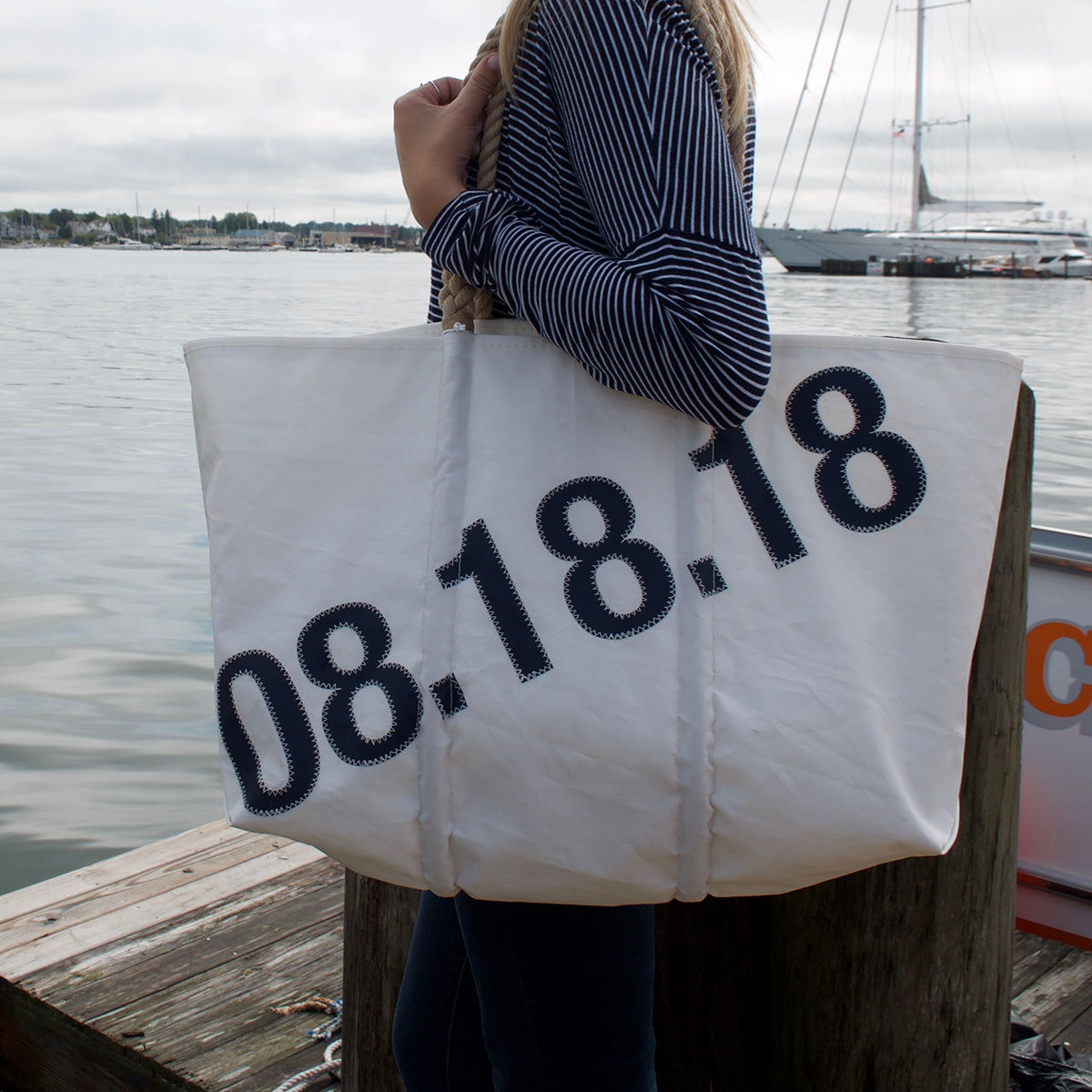 A person holding a spacious white Sea Bags Custom Date Tote featuring 08.18.18 in bold navy numbers, standing on a dock by the water with boats in the background—a perfect personalized tote bag and thoughtful anniversary gift. by Sea Bags