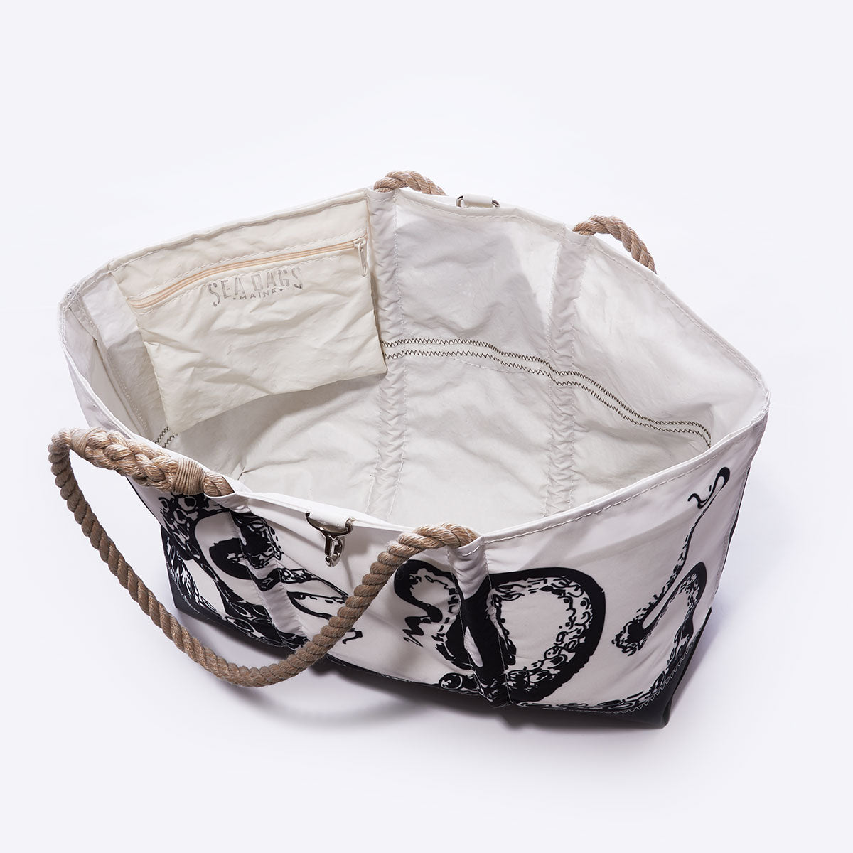 The Octopus Ogunquit Beach Tote by Sea Bags is a spacious white canvas bag, crafted from recycled sail cloth, featuring a black octopus print inspired by a vintage illustration. It includes sturdy rope handles, an interior pocket labeled with the Sea Bags brand, and is pictured open and empty against a plain white background. by Sea Bags