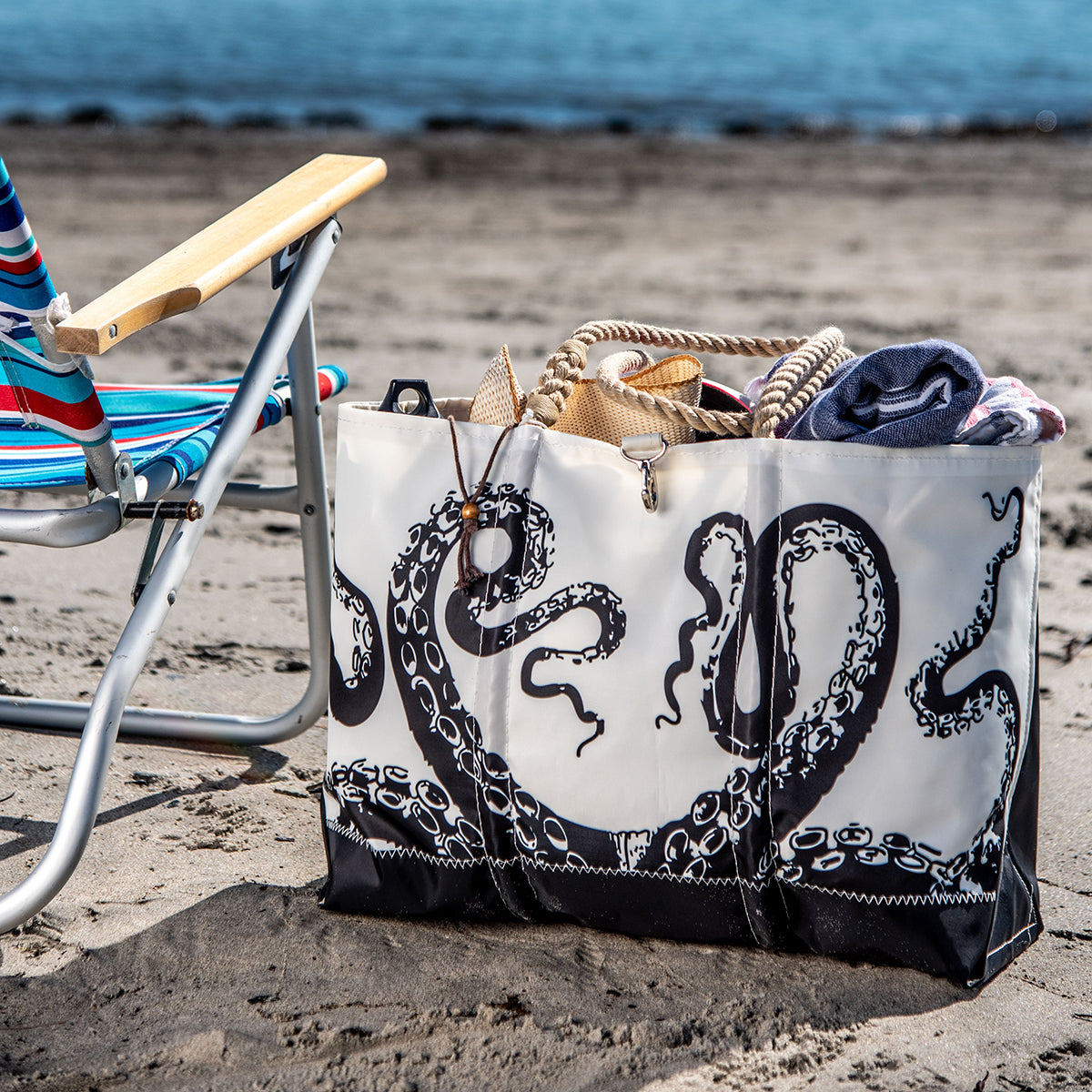 A Sea Bags Octopus Ogunquit Beach Tote sits on the sand near the water alongside a beach chair; crafted from recycled sail cloth and adorned with a vintage black octopus tentacle illustration, the spacious tote features sturdy rope handles and holds a hat and towel. by Sea Bags