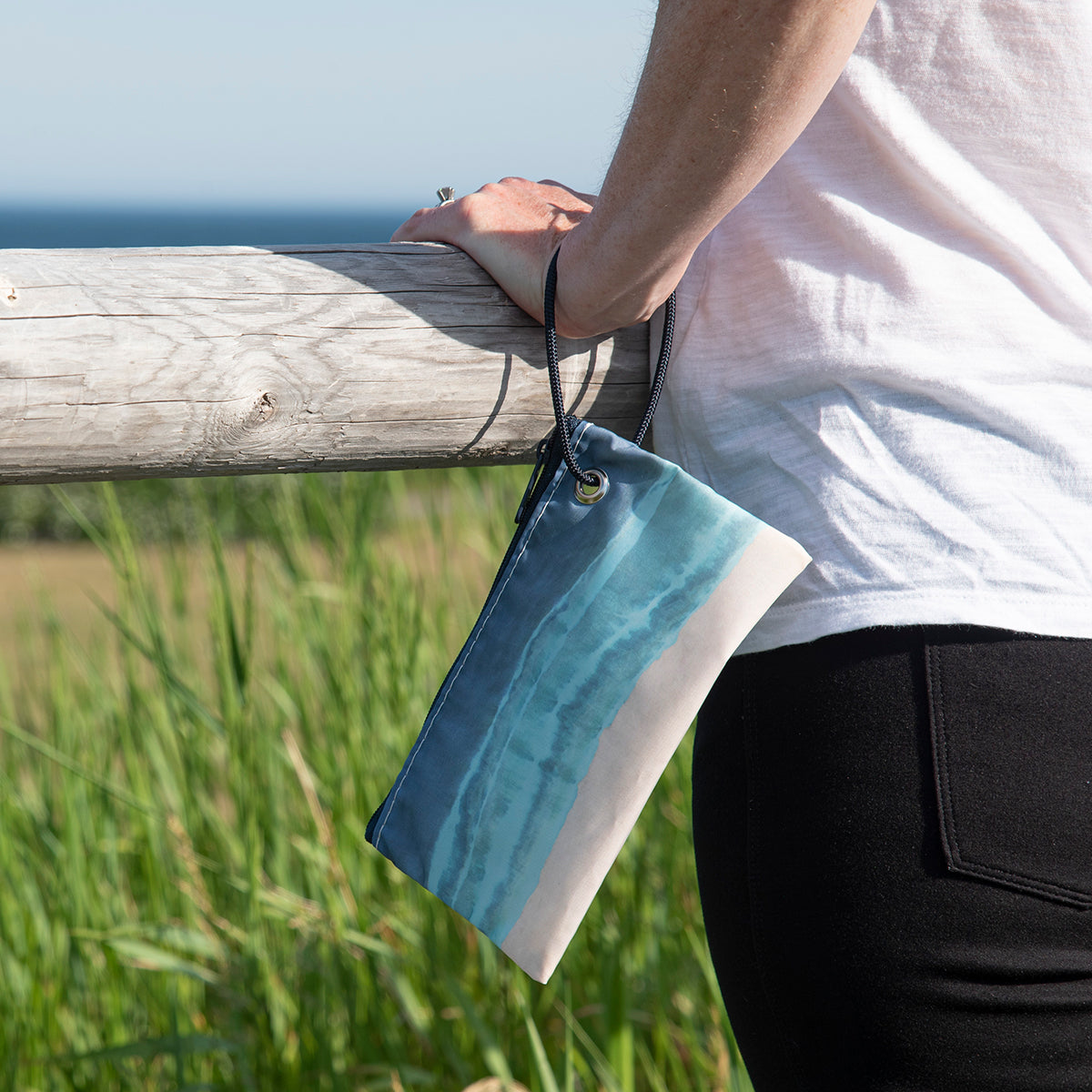A person stands by a wooden fence holding the Sea Bags Shoreline Tie Dye Wristlet, a small zippered pouch crafted from recycled sails and featuring a blue ocean and sandy beach design with a convenient wrist strap. by Sea Bags
