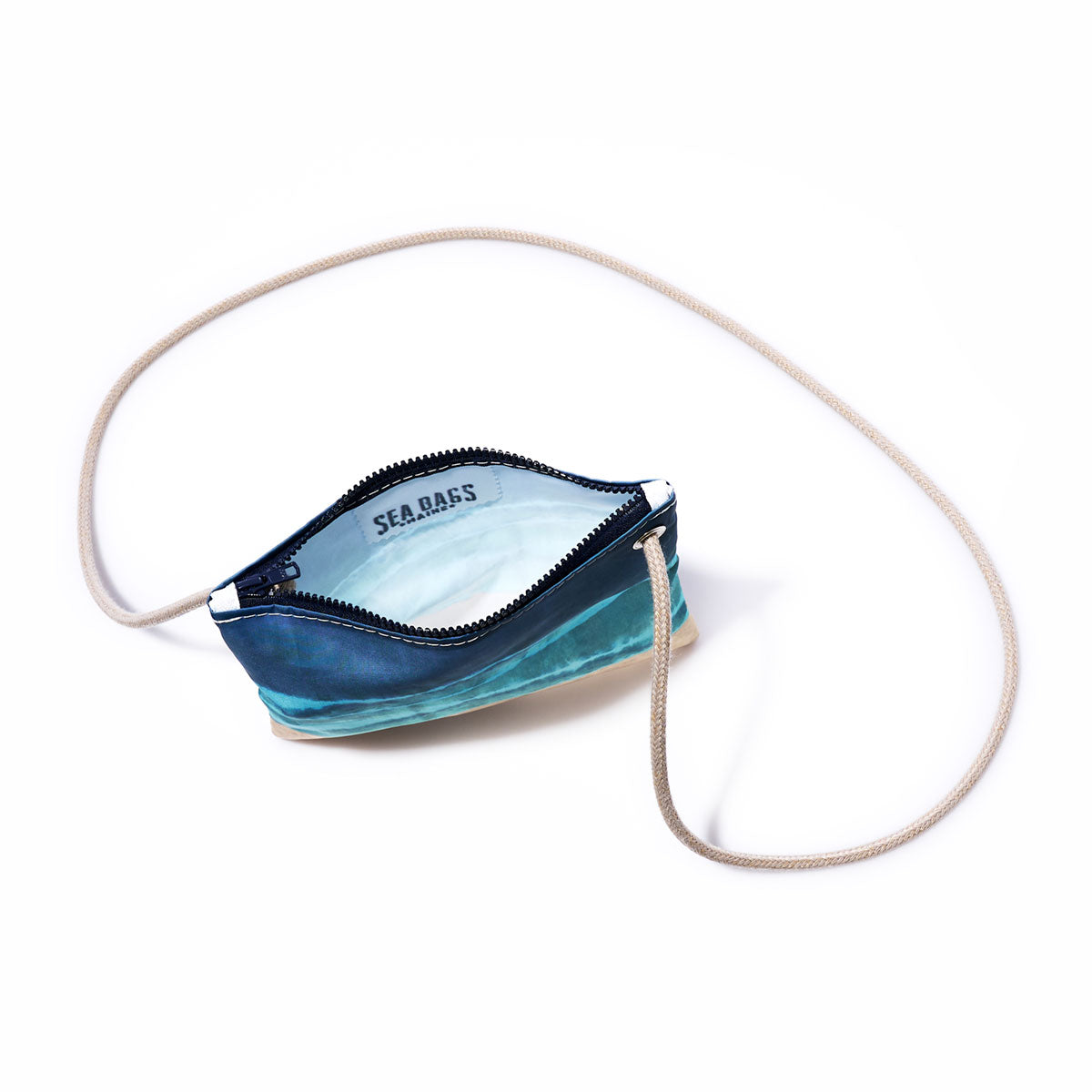 The Shoreline Tie Dye Slim Crossbody Bag by Sea Bags is a small zippered pouch made from recycled sail cloth, showcasing a blue-green wave design and finished with a cream-colored strap. With Sea Bags printed inside, this versatile accessory is ideal as a hands-free crossbody or an organizer for your favorite beach bag. Shown on a white background. by Sea Bags