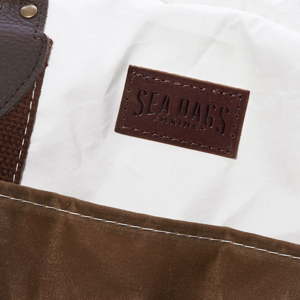 Close-up of the Tan Chebeague Weekender carry-on bag by Sea Bags, featuring a brown and white design crafted from recycled sail cloth. The detailed stitching enhances its durability, and a leather patch is stamped with the Sea Bags brand. by Sea Bags