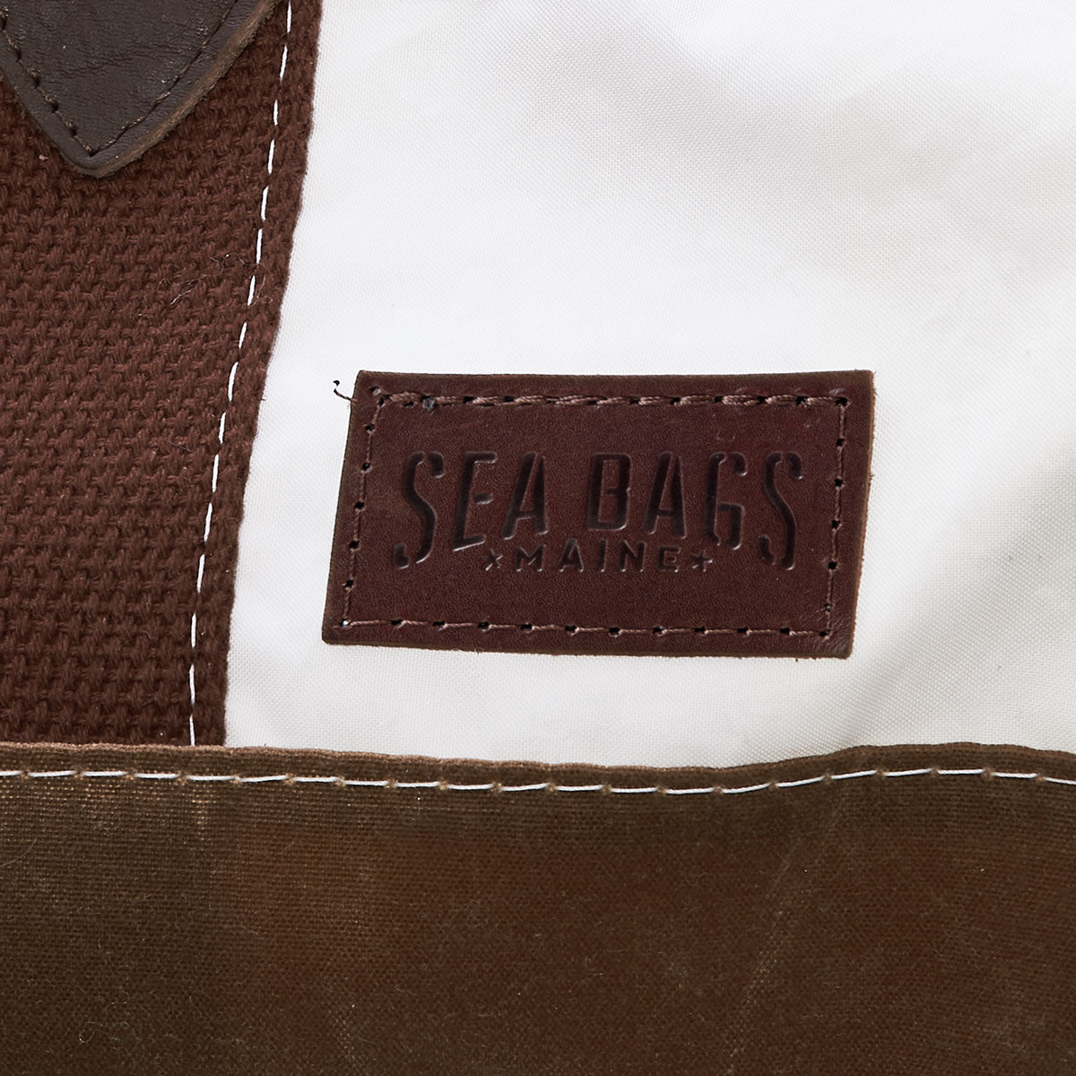 Close-up of the Tan Chebeague Duffel by Sea Bags, showing a brown rectangular patch with SEA BAGS MAINE text stitched onto fabric sections in brown and white, crafted from recycled sail cloth for an authentic maritime travel bag appearance. by Sea Bags