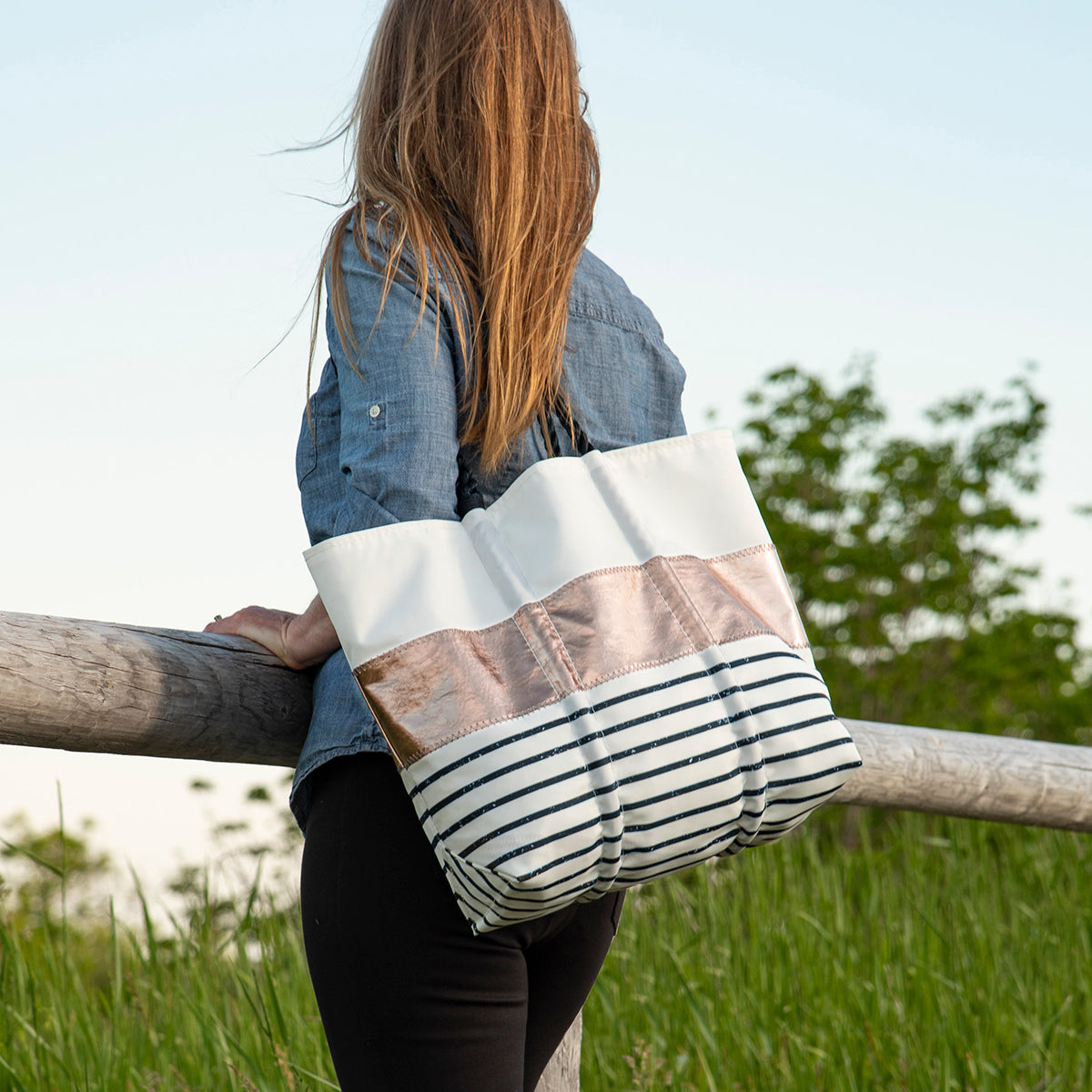 A woman with long brown hair stands by a wooden fence, wearing a denim shirt and carrying the Sea Bags Rose Gold-on-Navy Mariner Stripe Large Tote. by Sea Bags