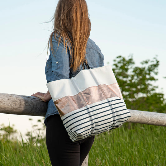 A woman with long brown hair stands by a wooden fence, wearing a denim shirt and carrying the Sea Bags Rose Gold-on-Navy Mariner Stripe Large Tote. by Sea Bags