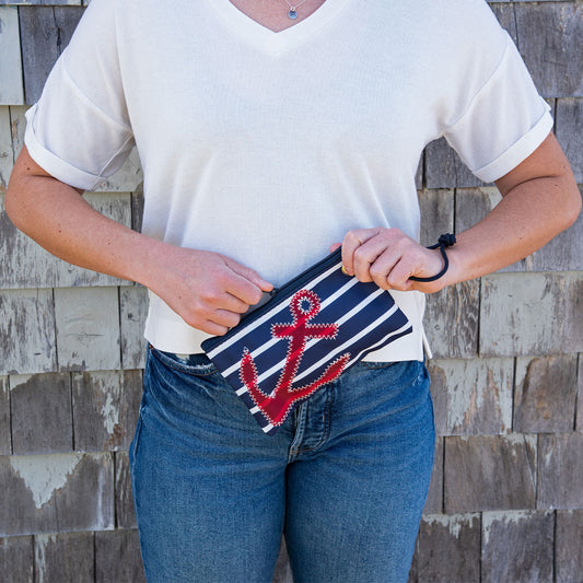 A person in a white shirt and blue jeans holds the Sea Bags Red Anchor on Navy Stripes Wristlet, crafted from recycled sails, in front of a weathered shingle wall. by Sea Bags