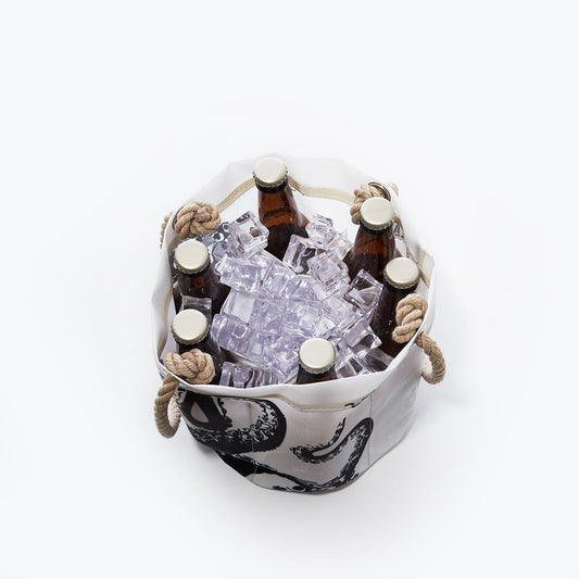 Six brown beer bottles with golden caps and clear ice cubes are nestled in a Sea Bags Octopus Beverage Bucket, crafted from recycled sail cloth with rope handles, viewed from above on a white background. by Sea Bags