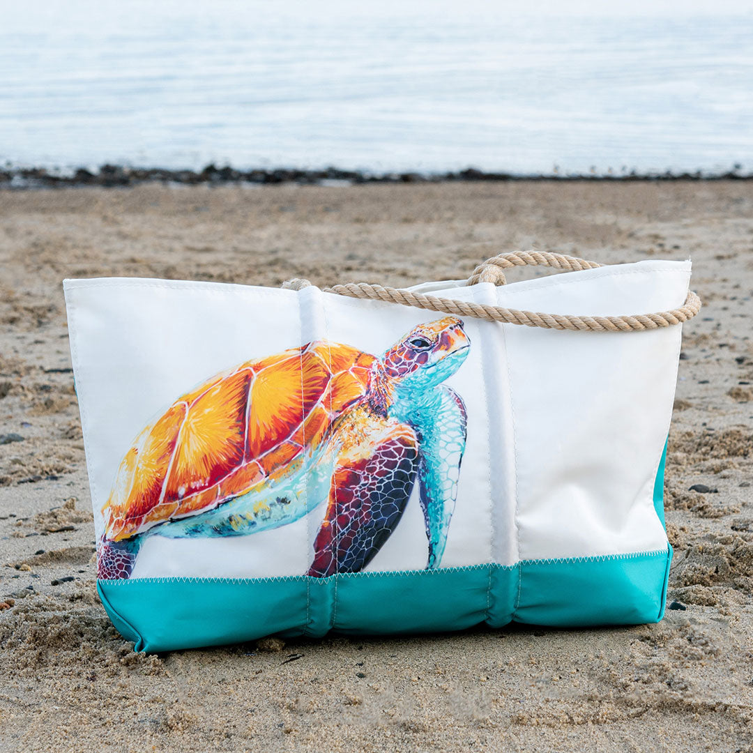 The Sea Bags Multicolor Sea Turtle Ogunquit Beach Tote, featuring a vibrant sea turtle design, rests on the sandy beach near the water. by Sea Bags