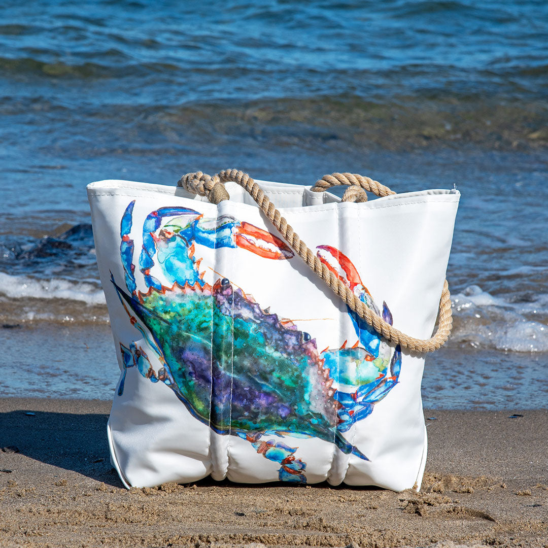 The Sea Bags Zip Top Watercolor Crab Medium Tote, crafted from recycled sail cloth by a Maine artist and featuring sturdy rope handles, showcases a colorful crab illustration as it sits on the sandy beach near the ocean. by Sea Bags