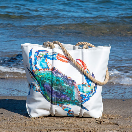 The Sea Bags Zip Top Watercolor Crab Medium Tote, crafted from recycled sail cloth by a Maine artist and featuring sturdy rope handles, showcases a colorful crab illustration as it sits on the sandy beach near the ocean. by Sea Bags