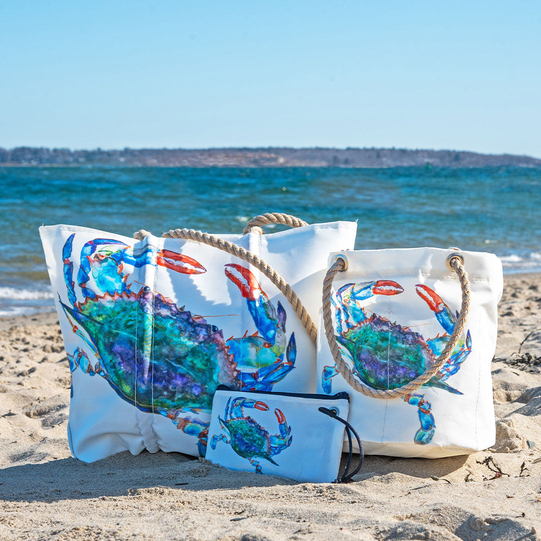Three white recycled sail bags with colorful blue crab designs and rope handles are displayed on a sandy beach near the ocean, with distant land visible across the water. Handcrafted by Maine artisans at Sea Bags, these unique bags capture coastal style—perfect for pairing with your favorite Watercolor Crab Wristlet. by Sea Bags