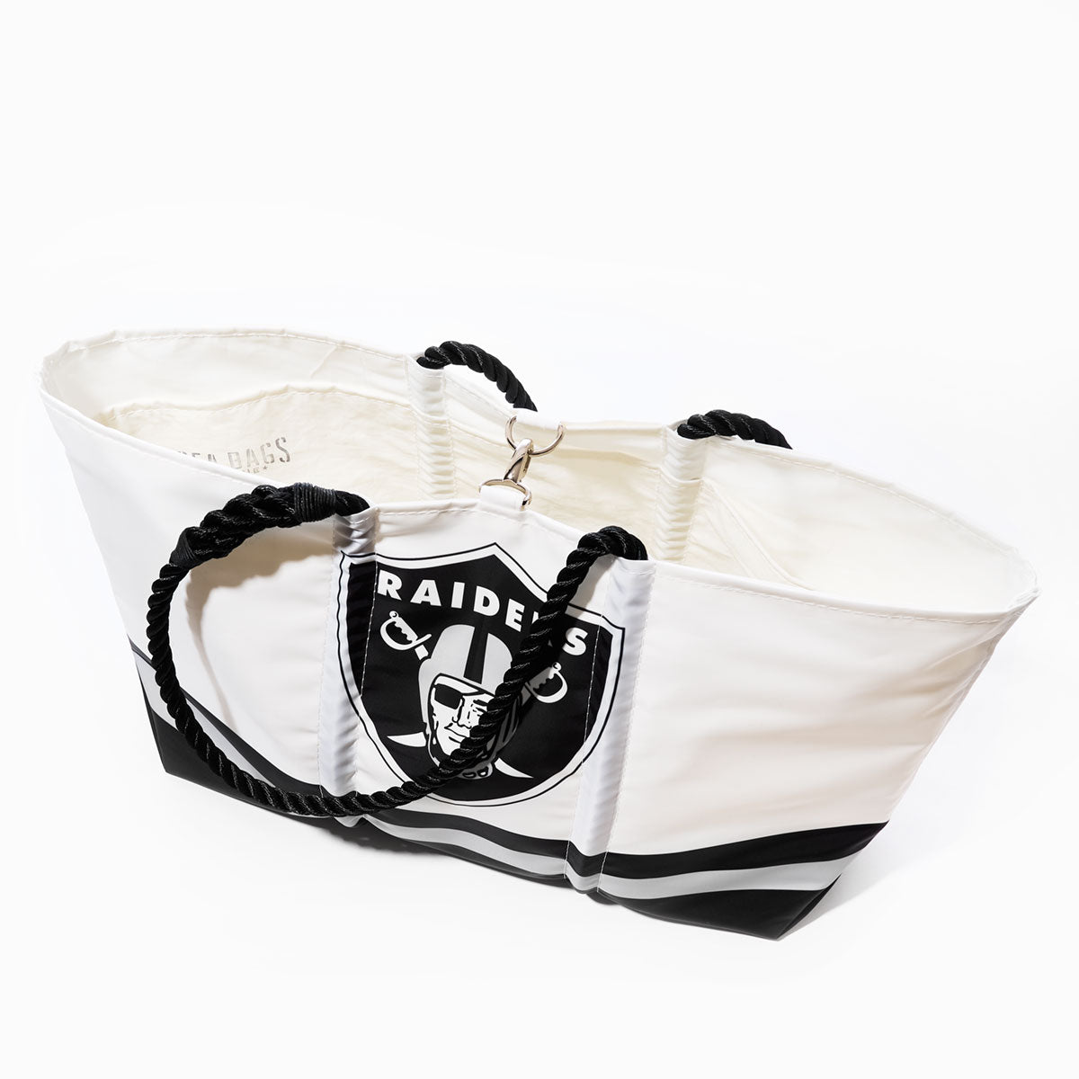 The Sea Bags Las Vegas Raiders Tailgate Tote is a large white bag made from recycled sail cloth, featuring sturdy black rope handles and a bold NFL logo on the front. The tote is displayed from an angled top view against a white background. by Sea Bags