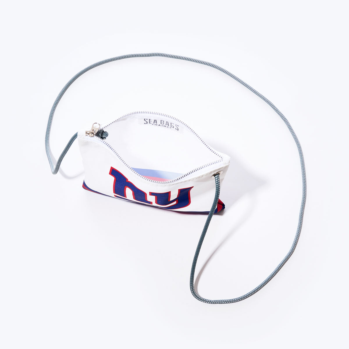 The Sea Bags New York Giants Slim Crossbody is a small white pouch made from recycled sail cloth, featuring a blue and red NY logo, gray cord strap, and zipper. Displayed open on a plain white background, this stylish and eco-friendly crossbody bag is the perfect accessory for NFL fans on the go. by Sea Bags