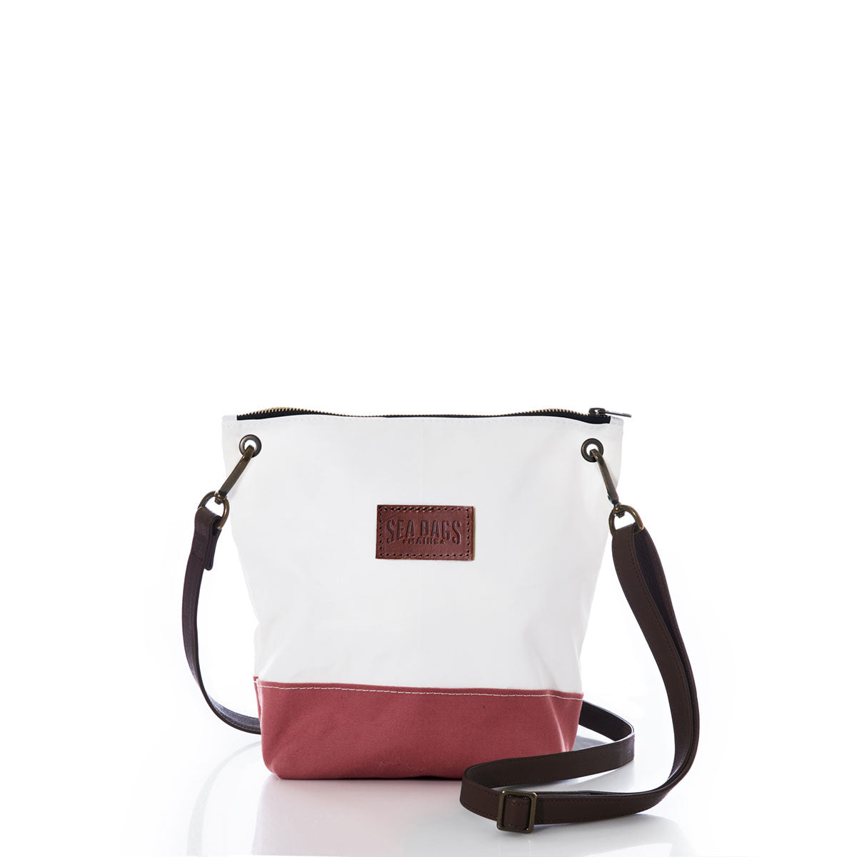 The Sea Bags Nantucket Red Chebeague Crossbody Bag features a two-tone design with a white upper made from recycled sail cloth and a Nantucket Red cotton canvas base. It is accented by a dark adjustable strap and a brown leather Sea Bags patch on the front, all displayed against a plain white background. by Sea Bags