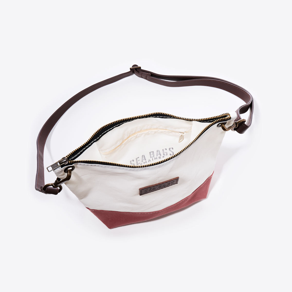 The Nantucket Red Chebeague Crossbody Bag by Sea Bags features a brown shoulder strap, a white upper section made from recycled sail cloth, and a Nantucket Red cotton canvas base; pictured unzipped to reveal its light-colored interior with a SEA BAGS label. by Sea Bags