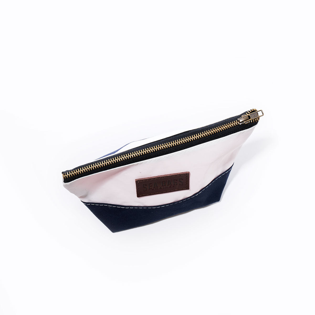 The Chebeague Cosmetic Bag by Sea Bags is a small zippered pouch featuring a white upper and dark blue lower section, expertly crafted from recycled sailcloth, canvas, and leather. It includes a brass zipper and a brown patch label on one side—making it the perfect stylish cosmetic bag. Shown here against a plain white background. by Sea Bags