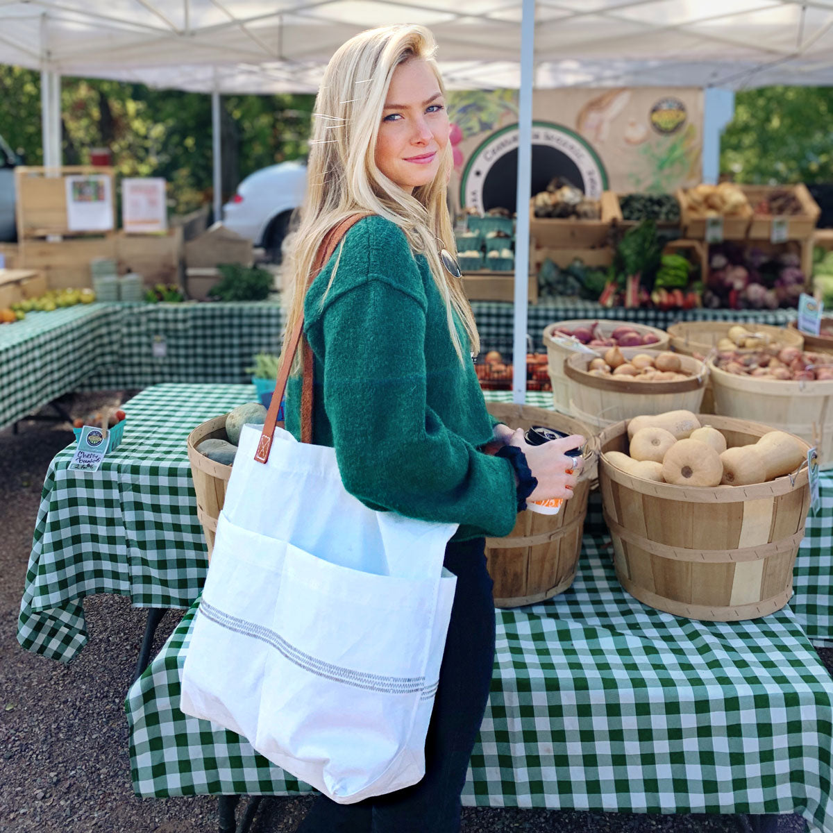 A woman with long blonde hair stands by produce baskets at an outdoor farmers market, wearing a green sweater and carrying the Sea Bags Market Tote with white leather handles; green checked tablecloths cover the tables. by Sea Bags