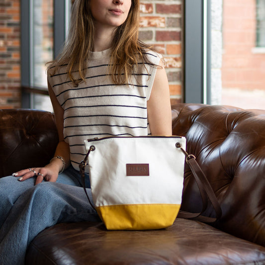 A woman sits on a brown leather couch near a window with brick walls in the background, next to the Sea Bags Golden Yellow Chebeague Crossbody Bag, made from white and yellow recycled sailcloth with a leather patch. by Sea Bags