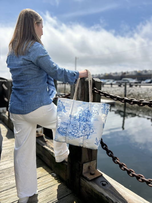 A woman wearing a denim shirt and white pants stands on a dock along the picturesque Maine coast, resting one foot on a wooden post and holding the Sea Bags Maine Fishing Village Medium Tote, crafted from recycled sail cloth with sturdy rope handles. by Sea Bags