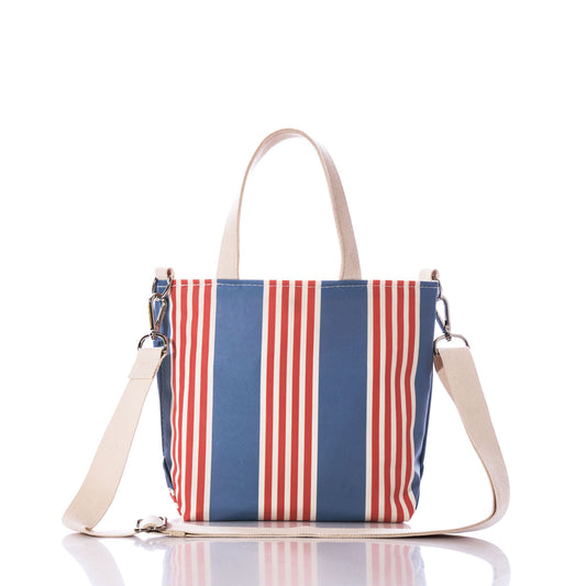The French Stripe Crossbody Tote by Sea Bags is a small recycled sailcloth bag showcasing blue and red vertical stripes in a classic French stripe design. It features two short cream handles and a detachable cream shoulder strap, photographed on a white background. by Sea Bags