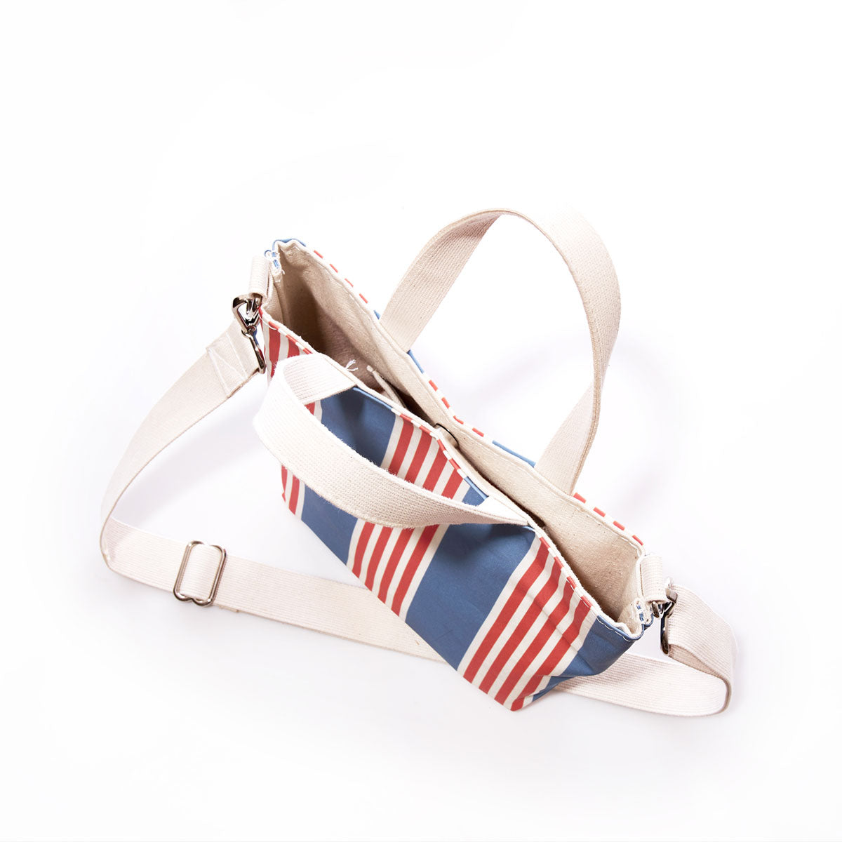 The Sea Bags French Stripe Crossbody Tote, crafted from recycled sailcloth, is displayed on a white background. It showcases a French stripe design with blue and red vertical stripes, complemented by a beige adjustable strap and an open main compartment. This tote-crossbody hybrid effortlessly blends style and functionality for everyday use. by Sea Bags
