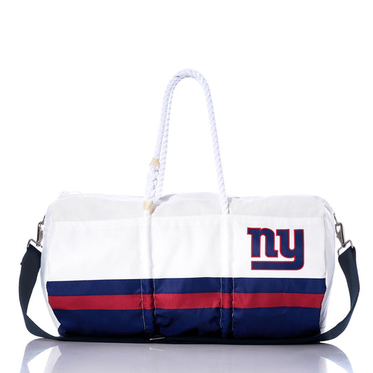 New York Giants Duffel by Sea Bags, made from recycled sail cloth with white and blue panels, red stripes, a rope handle, black strap, and NY logo on the side—shown on a reflective surface. by Sea Bags