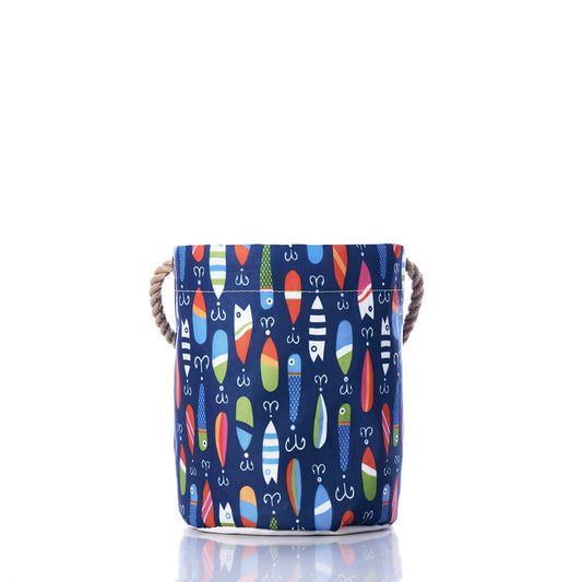 The Kate Nelligan Lures Bucket Bag by Sea Bags features a blue fabric design with a multicolored surfboard pattern, sturdy rope handles on each side, and coastal prints that evoke a nautical feel, all set against a crisp white background. by Sea Bags