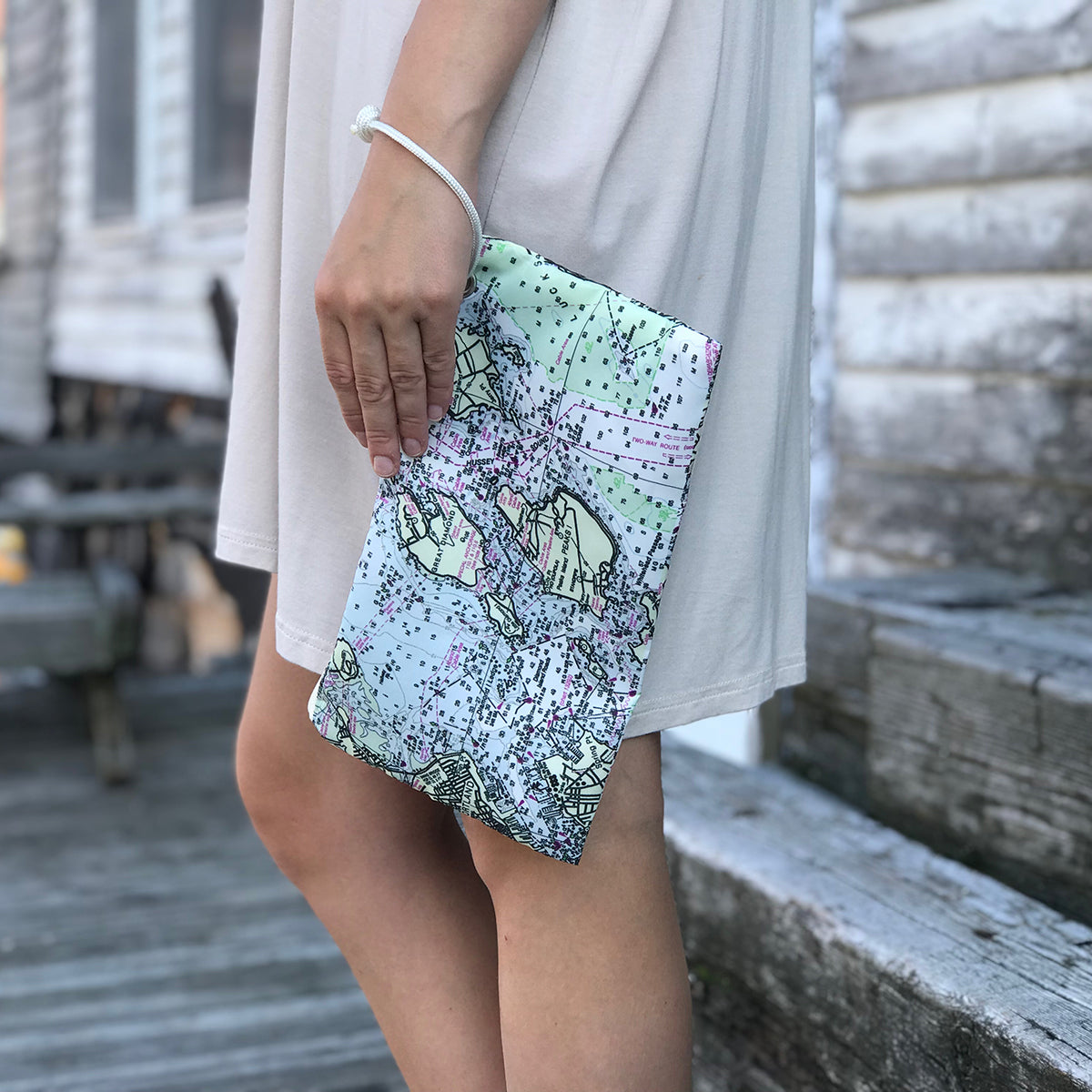 A person in a light dress stands outside holding the Sea Bags Casco Bay Nautical Chart Wristlet—a pastel-colored zip-top accessory crafted from recycled sail cloth and featuring a map-inspired design reminiscent of Casco Bay; the background shows weathered wooden steps and siding. by Sea Bags