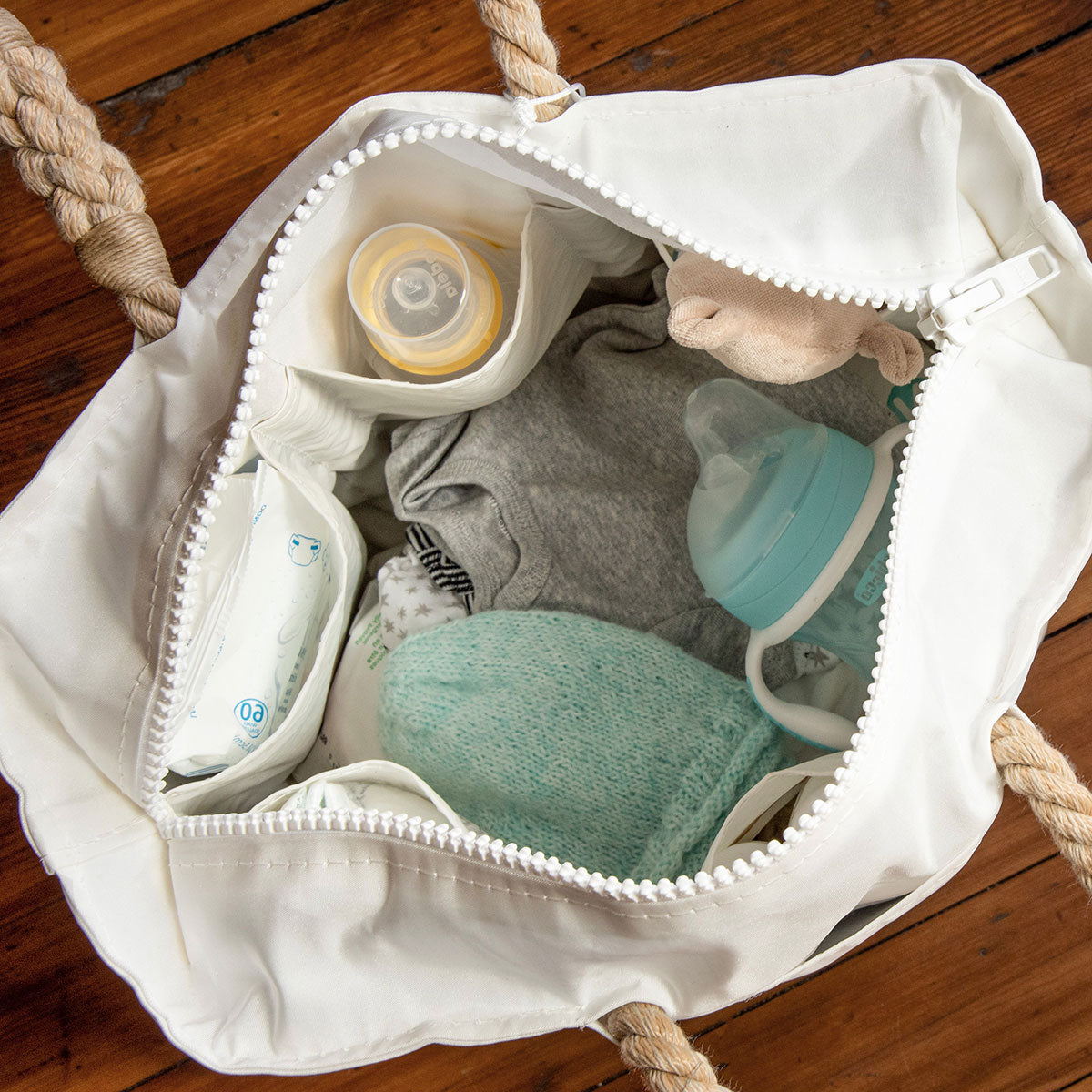 The Sea Bags Custom Design Diaper Bag-White, shown open on a wooden floor, holds a baby bottle, pacifier, wipes, gray shirt, plush toy, diaper, and teal washcloth. by Sea Bags