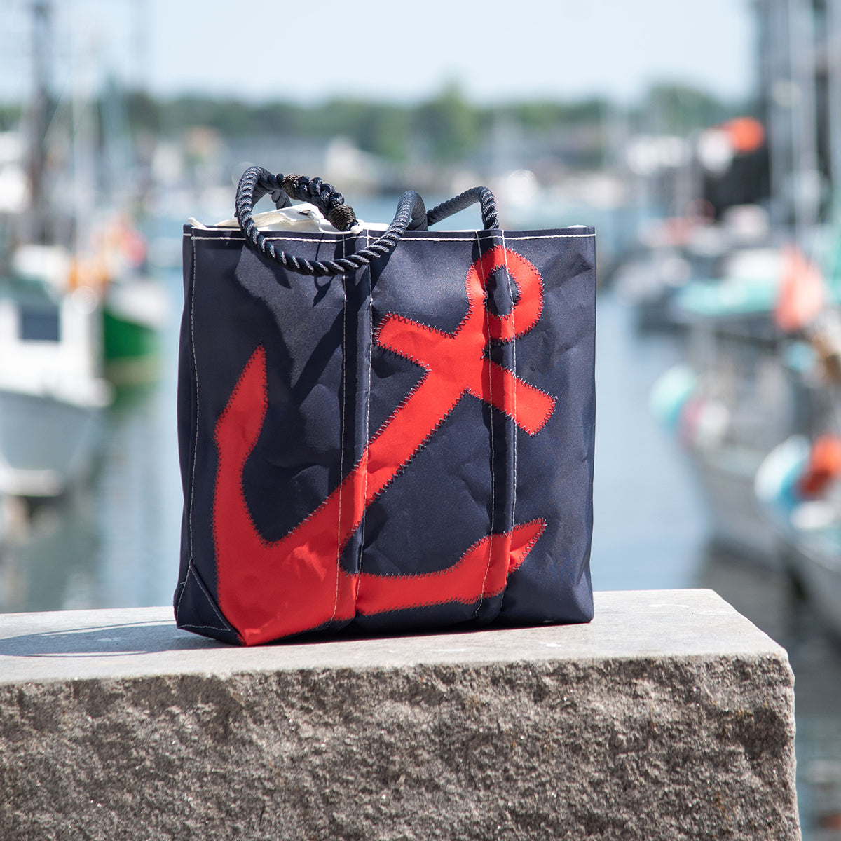 Sea Bags Custom Design Medium Tote in navy blue reclaimed sailcloth, featuring a bold red anchor motif, shown on a stone ledge overlooking a marina with boats in the background. by Sea Bags