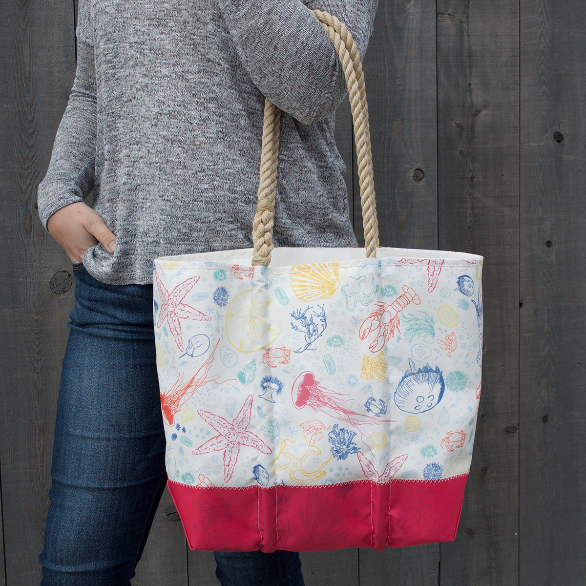 A person holding the Sea Bags Custom Design Medium Tote, which has sturdy rope handles, a sea creature print, and a solid pink bottom, stands in front of a wooden background. by Sea Bags