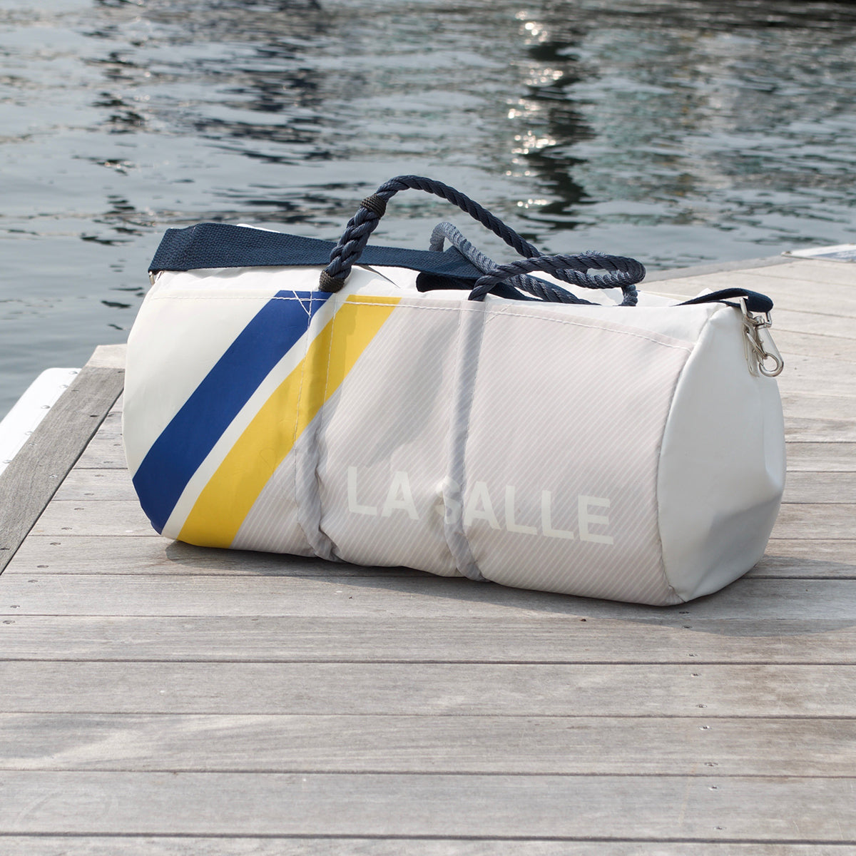A Sea Bags Custom Design Duffel featuring blue and yellow stripes and LA GALE text rests on a wooden dock by the water, making it the perfect personalized travel companion for your next adventure. by Sea Bags