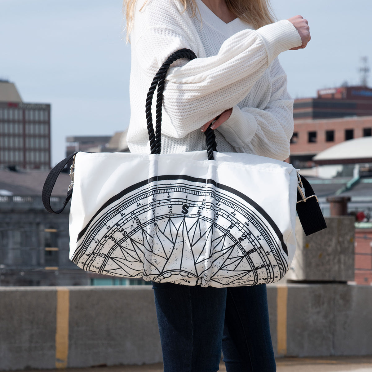 A person in a white sweater stands outdoors in an urban setting, holding the Sea Bags Custom Design Duffel, featuring a bold black compass graphic. by Sea Bags