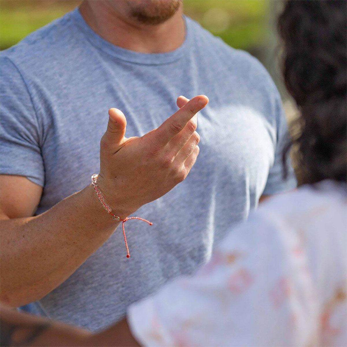A person in a gray t-shirt gestures with their hand, wearing a 4ocean Beaded Bracelet - Coral Reef by 4ocean, demonstrating their commitment to a clean ocean. Another person in a patterned shirt is partially visible in the foreground. by Sea Bags