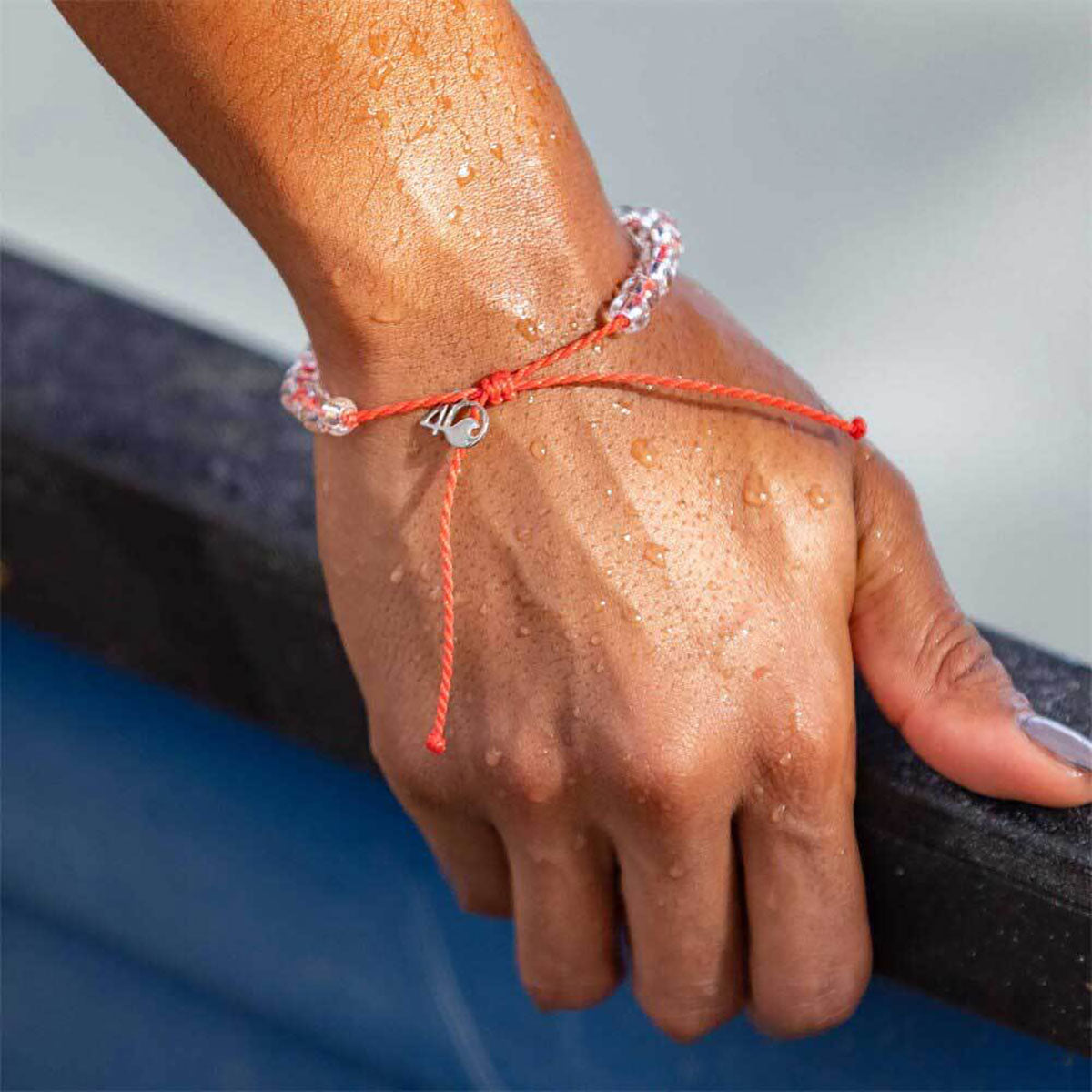 A close-up of a wet hand resting on a blue surface, wearing a red string bracelet with a silver charm and a beaded bracelet, alongside the 4ocean Beaded Bracelet - Coral Reef by 4ocean, an eco-friendly accessory that represents a commitment to clean ocean initiatives. by Sea Bags
