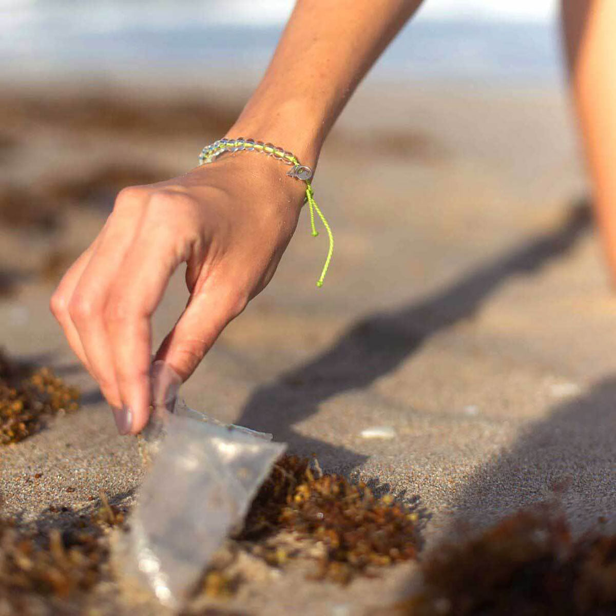 Wearing the 4ocean Beaded Bracelet - Sea Turtle by 4ocean, a person picks up a piece of plastic trash from the sandy beach, surrounded by seaweed—a symbol of ocean conservation and sustainable jewelry. by Sea Bags